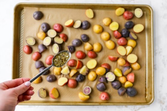 Sliced potatoes on a sheet pan lined with parchment paper. A hand is holding a spoon, drizzling Lemon Garlic Sauce over the potatoes.