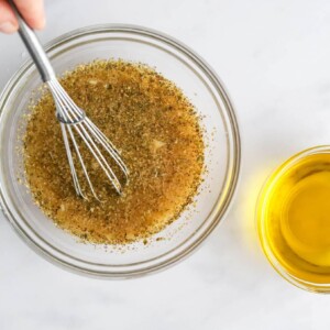 Apple cider vinegar, lemon juice, water, dijon mustard, honey, dried oregano, garlic, sea salt and black pepper whisked together in a bowl. A small bowl of olive oil is nearby.