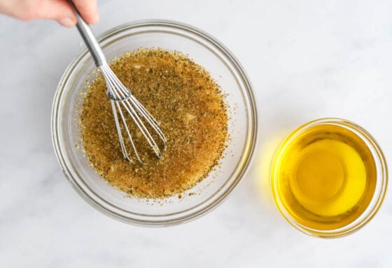 Apple cider vinegar, lemon juice, water, dijon mustard, honey, dried oregano, garlic, sea salt and black pepper whisked together in a bowl. A small bowl of olive oil is nearby.