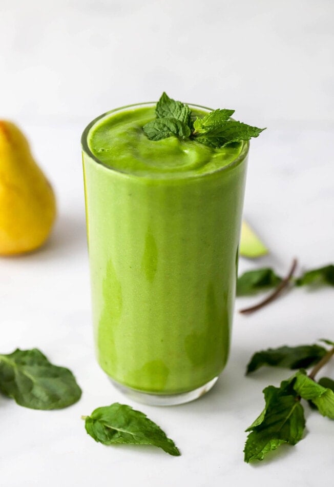 A glass filled with pear smoothie. Fresh mint tops the glass and is scattered around.