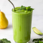 A glass filled with pear smoothie. Fresh mint tops the glass and is scattered around. A straw sticks out of the glass.