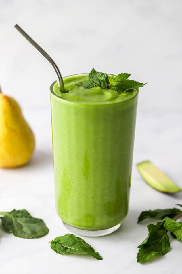 A glass filled with pear smoothie. Fresh mint tops the glass and is scattered around. A straw sticks out of the glass.