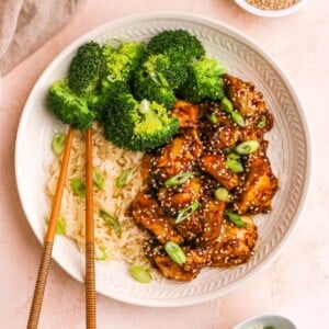 An overhead view of a plate of sesame chicken served with rice and broccoli. Chopsticks lay across the plate.