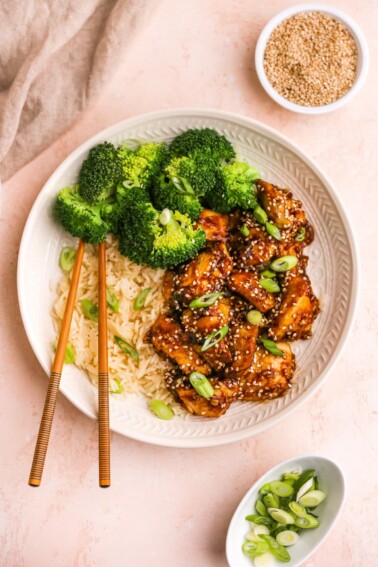 An overhead view of a plate of sesame chicken served with rice and broccoli. Chopsticks lay across the plate.