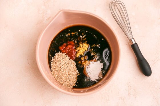 Ingredients for sesame chicken sauce in a mixing bowl: tamari, rice vinegar, water, sesame oil, arrowroot powder, coconut sugar, garlic, fresh ginger, sambal oelek, sea salt and sesame seeds.