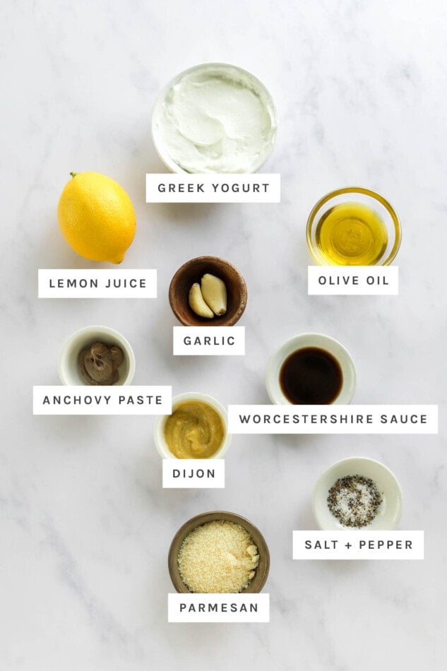 Ingredients measured out to make healthy caesar dressing: Greek yogurt, olive oil, lemon juice, garlic, anchovy paste, Worcestershire sauce, dijon mustard, salt, pepper and parmesan.