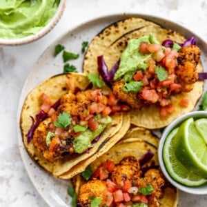 Three cauliflower tacos on a plate with a ramekin of lime wedges. The tacos are topped with avocado crema and fresh cilantro.