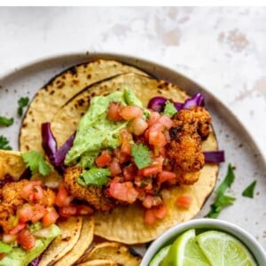 Closeup of 3 tacos on a plate with a ramekin of limes. The tacos are topped with fresh cilantro and avocado crema.