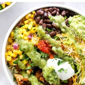 A chicken burrito bowl with corn salad, black beans, guacamole, shredded cheese, lime slices, greek yogurt and salsa served over cilantro lime rice. It has been drizzled with a tomatillo avocado dressing.
