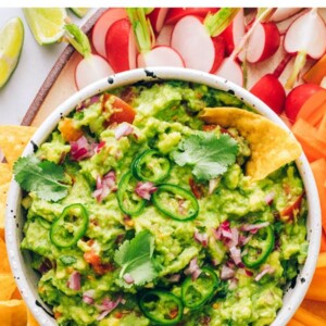 A serving bowl of guacamole surrounded by carrots, radishes, chips, celery, cucumber slices, orange pepper and carrots for dipping.