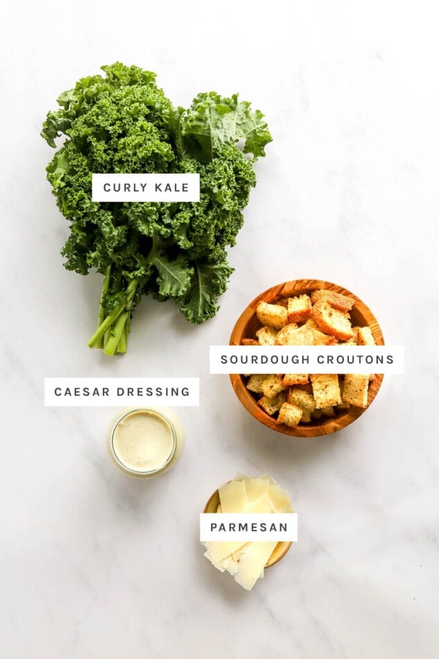 Ingredients measured out to make a kale caesar salad: curly kale, sourdough croutons, caesar dressing and parmesan.