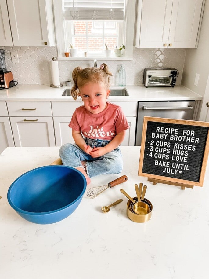 Toddler girl in a big sister shirt with recipe for baby brother sign.