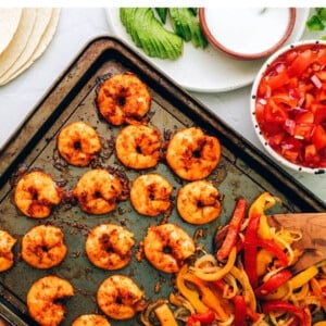 Cooked veggies and shrimp on a sheet pan. Fajitas accoutrements surround the sheet pan.