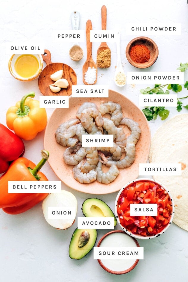 Ingredients measured out to make shrimp fajitas: pepper, cumin, chili powder, onion powder, olive oil, sea salt, garlic, cilantro, shrimp, tortillas, bell peppers, onion, avocado, salsa and sour cream.