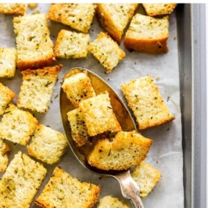 Sourdough croutons on a baking sheet lined with parchment paper. A spoon is lifting a few croutons off the baking sheet.