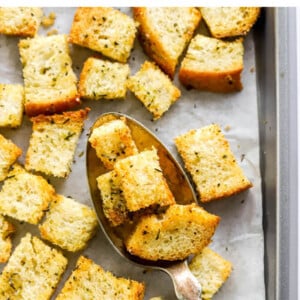 Sourdough croutons on a baking sheet lined with parchment paper. A spoon is lifting a few croutons off the baking sheet.
