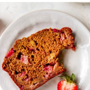 A slice of strawberry banana bread with a bite taken out of it, resting on a plate with a halved strawberry.