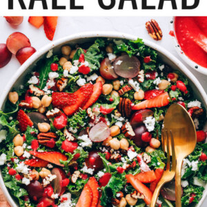 Large bowl with a kale salad topped with strawberries, grapes, feta, pecans and chickpeas. Two serving utensils are in the bowl.