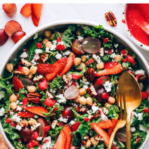 Large bowl with a kale salad topped with strawberries, grapes, feta, pecans and chickpeas. Two serving utensils are in the bowl.