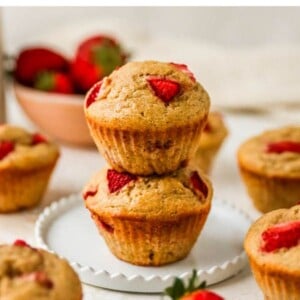 Two strawberry yogurt muffins stacked on top of each other on a plate. Other muffins are scattered around the plate.
