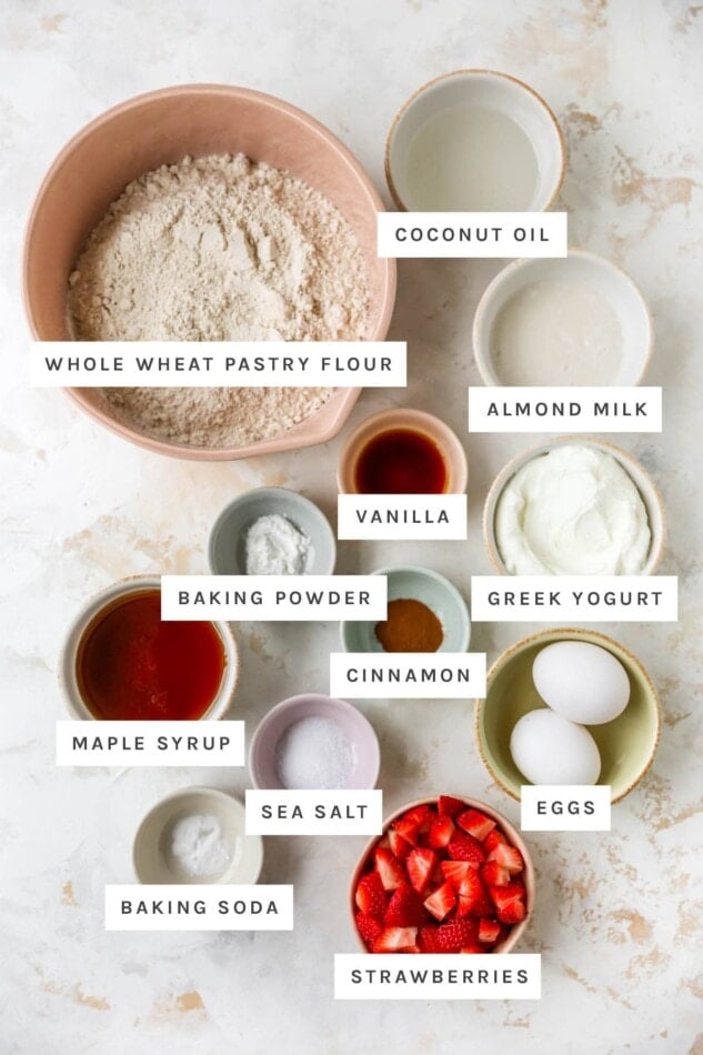 Ingredients measured out to make strawberry yogurt muffins: whole wheat pastry flour, coconut oil, almond milk, vanilla, Greek yogurt, vanilla, baking powder, cinnamon, eggs, maple syrup, sea salt, baking soda and strawberries.
