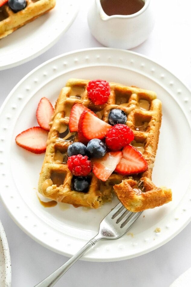 An almond flour waffle on a white plate topped with strawberries, blueberries and raspberries and a drizzle of maple syrup. A fork has a piece of waffle on it and is resting on the plate.