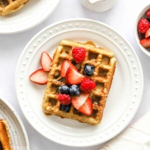 An almond flour waffles on a white plate. It has been topped with various berries and a drizzle of maple syrup.