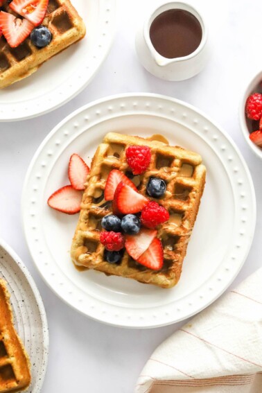 An almond flour waffles on a white plate. It has been topped with various berries and a drizzle of maple syrup.