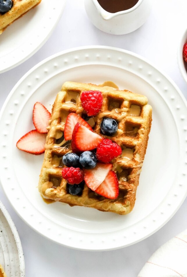 An almond flour waffles on a white plate. It has been topped with various berries and a drizzle of maple syrup.