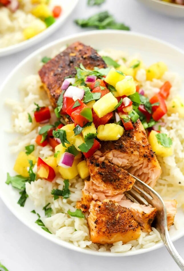 A filet of blackened salmon served over a bed of rice topped with pineapple salsa. A fork is removing a bite from the filet.