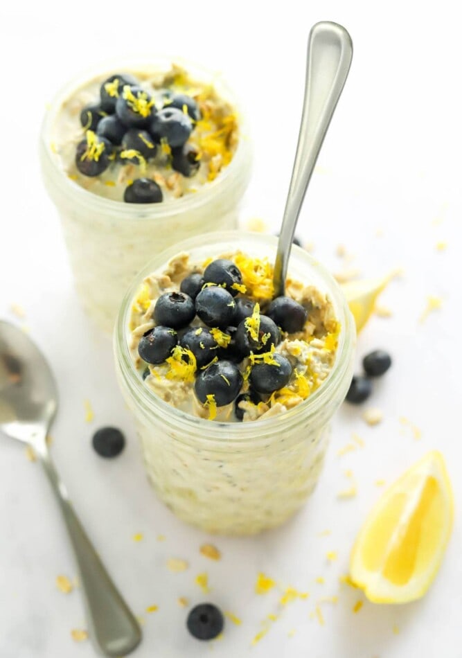 Two jars of blueberry lemon overnight oats topped with extra blueberries and lemon zest. A spoon is resting in the front jar.