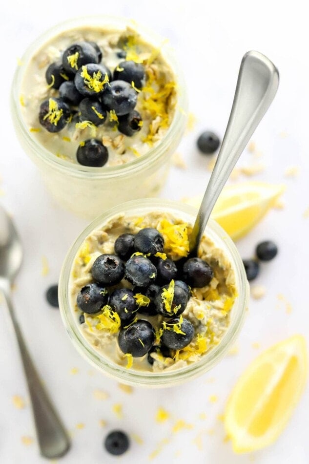 Overhead view of overnight oats in a mason jars topped with blueberries and lemon zest. A spoon is sticking out of one of the jars.
