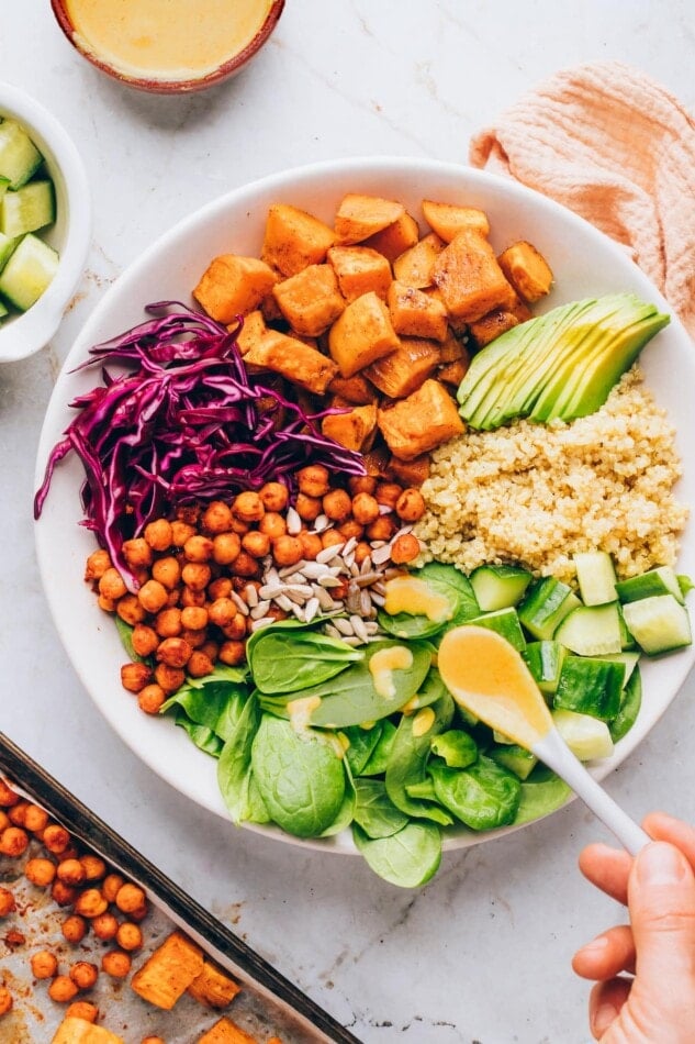 A bowl containing sweet potatoes, spinach, sunflower seeds, shredded red cabbage, chickpeas, cucumbers, quinoa and avocado. A small spoon is drizzling dressing over the bowl.