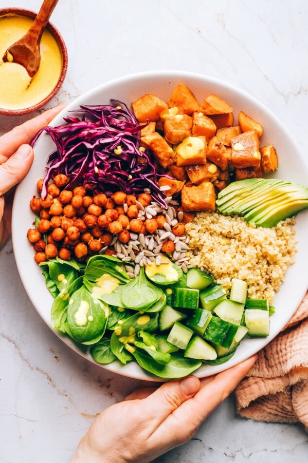 Hands holding a bowl containing sweet potatoes, spinach, sunflower seeds, shredded red cabbage, chickpeas, cucumber, quinoa and avocado.