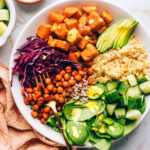 A buddha bowl with sweet potatoes, spinach, sunflower seeds, shredded red cabbage, chickpeas, cucumbers, quinoa and avocado. A fork rests in the bowl.