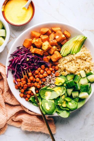 A buddha bowl with sweet potatoes, spinach, sunflower seeds, shredded red cabbage, chickpeas, cucumbers, quinoa and avocado. A fork rests in the bowl.