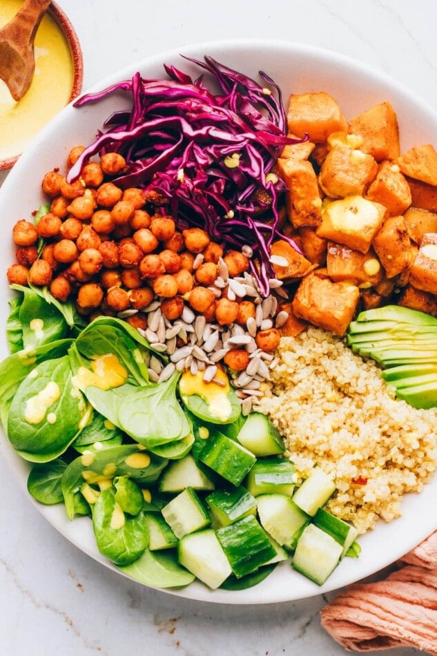 Closeup view of a bowl containing sweet potatoes, spinach, sunflower seeds, shredded red cabbage, chickpeas, cucumbers, quinoa and avocado