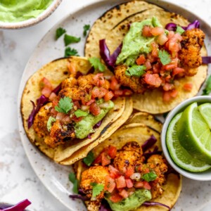 Three cauliflower tacos on a plate. A small ramekin with limes is also on the plate.