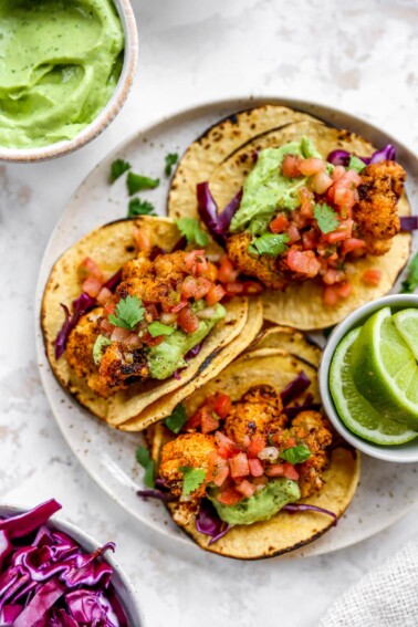 Three cauliflower tacos on a plate. A small ramekin with limes is also on the plate.