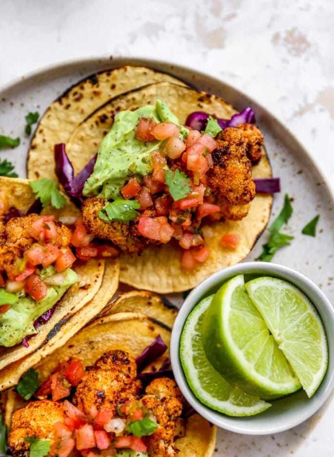 Closeup of 3 tacos on a plate with a ramekin of limes. The tacos are topped with fresh cilantro and avocado crema.