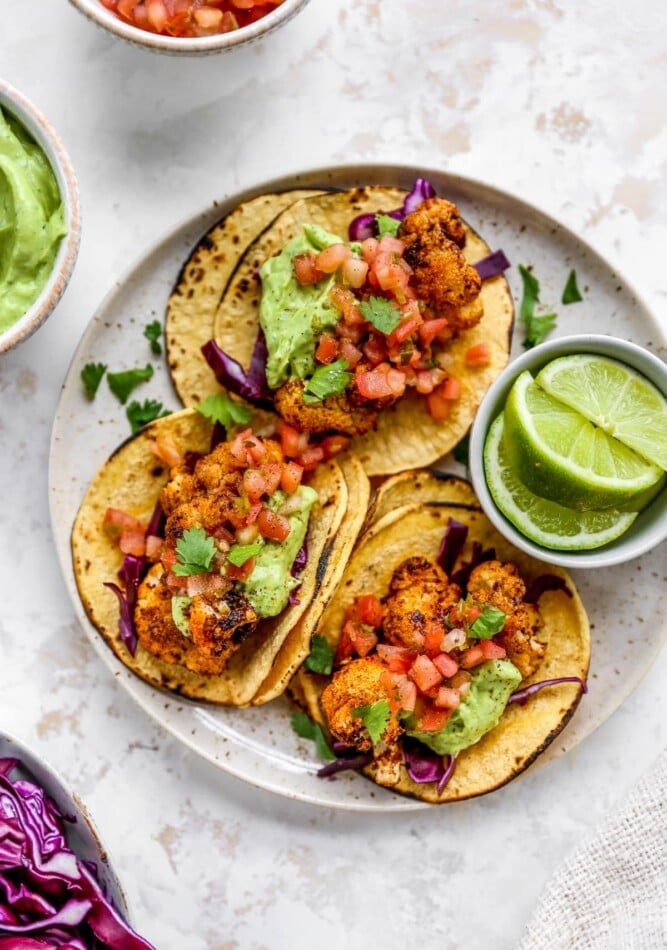 Three cauliflower tacos on a plate with a ramekin of lime wedges. The tacos are topped with avocado crema and fresh cilantro.