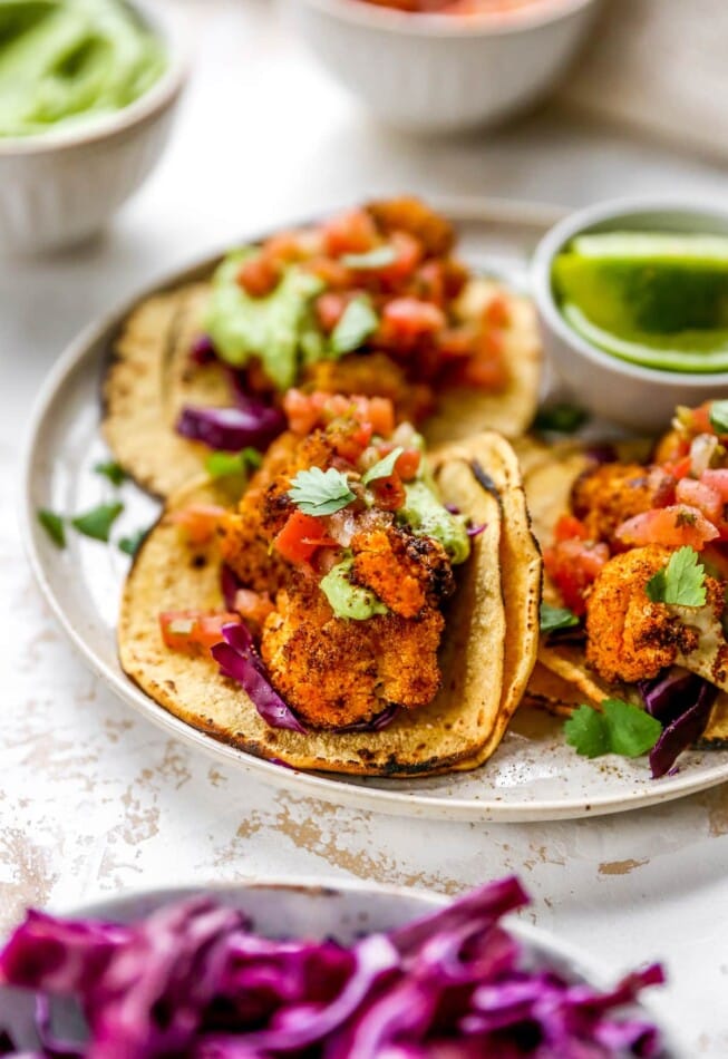 A cauliflower taco topped with avocado crema and fresh cilantro in focus on plate. There are 2 other tacos on the plate.