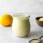 A small tulip jar containing caesar salad dressing. A small spoon rests next to the jar.