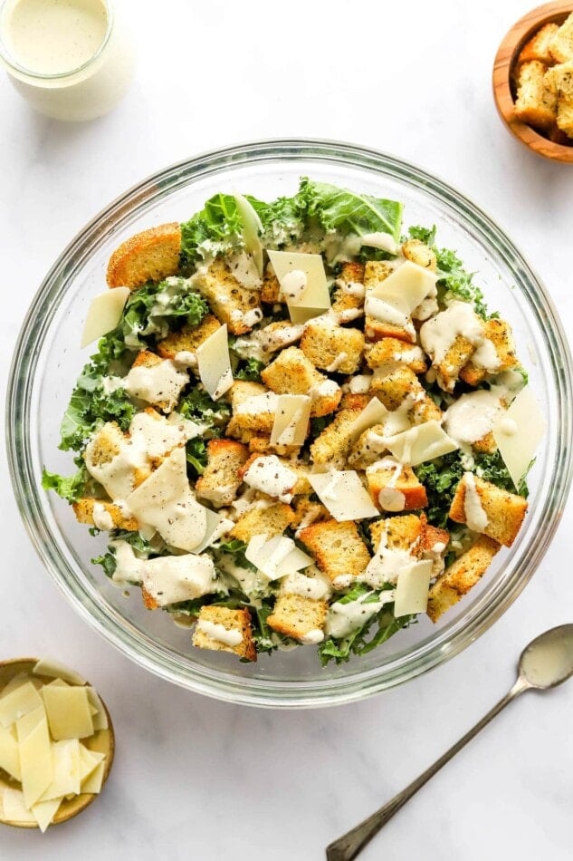 Overhead view of a large serving bowl containing kale salad topped with sourdough croutons, parmesan flakes and healthy caesar dressing. A spoon with dressing rests to the side of the bowl.