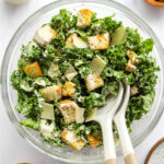 A serving bowl containing kale caesar salad. Two serving spoons rest in the bowl.