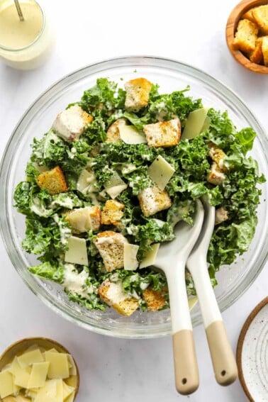 A serving bowl containing kale caesar salad. Two serving spoons rest in the bowl.