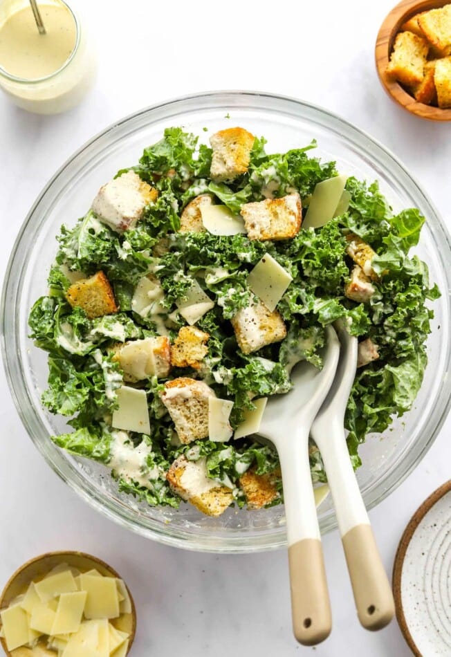 A serving bowl containing kale caesar salad. Two serving spoons rest in the bowl.