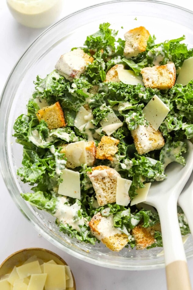 A serving bowl containing kale caesar salad. Two serving spoons rest in the bowl.