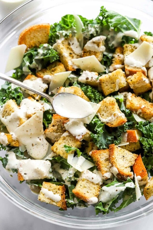 Drizzling healthy caesar dressing onto kale salad topped with sourdough croutons and parmesan flakes.