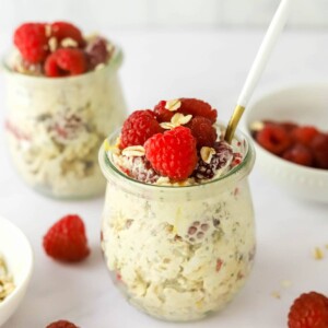 A jar of raspberry lemon overnight oats topped with extra raspberries and lemon zest. A spoon rests in the jar. A second jar is in the background.